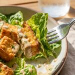 A close-up of a fork lifting a piece of crispy tofu with dressing and greens.