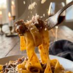 A close-up of a fork twirling pappardelle pasta with meaty Bolognese sauce, with grated cheese falling from above.