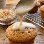 A close-up of a spoon pouring creamy milk over a poked cupcake so it can soak into the sponge