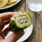 A close-up of a hand holding a pickle boat with a bite taken out, showing the creamy tuna inside.