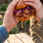 A close-up of two hands holding a messy BBQ jackfruit sandwich with slaw, ready to take a bite.