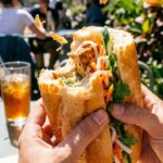 Two hands holding a partially eaten vegan banh mi sandwich, showing the layers of tofu and pickled veggies with crumbs falling from the crusty bread.