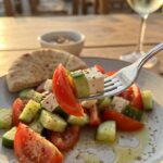 A close-up of a fork lifting a bite of tomato, cucumber, and vegan feta from a Greek salad, dripping with dressing.
