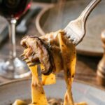 A close-up of a fork twirling a bite of creamy mushroom stroganoff pasta, with steam rising and wine in the background.
