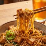 A close-up of chopsticks lifting glossy pad thai noodles with tofu and peanuts, with a squeeze of lime juice falling on them.