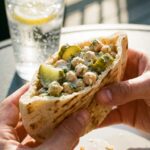 A close-up of hands holding a stuffed chickpea and pickle pita pocket, ready to take a bite.
