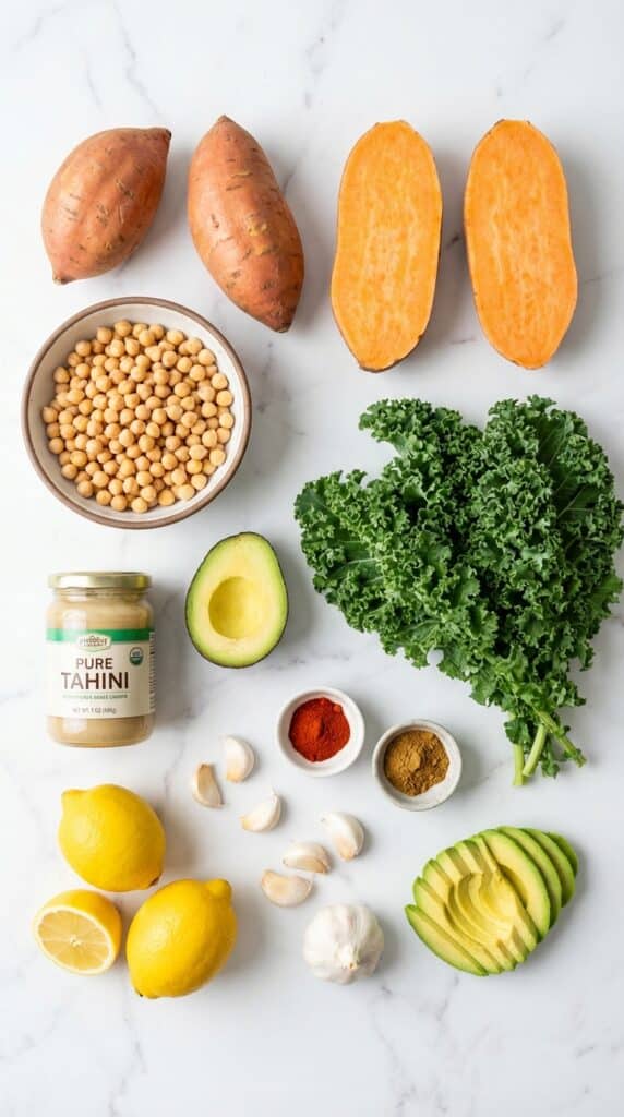 A flat lay showing raw sweet potatoes, chickpeas, kale, tahini, lemons, avocado, and spices on a white marble board.