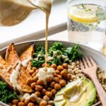 A close-up of thick, creamy tahini dressing being poured from a small pitcher over a bowl of sweet potatoes, chickpeas, and avocado.
