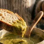 A close-up of a piece of crusty bread dipping into a hot bowl of thick split pea soup, with steam rising.