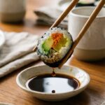 A close-up of chopsticks dipping a piece of vegetable sushi into a small dish of soy sauce.