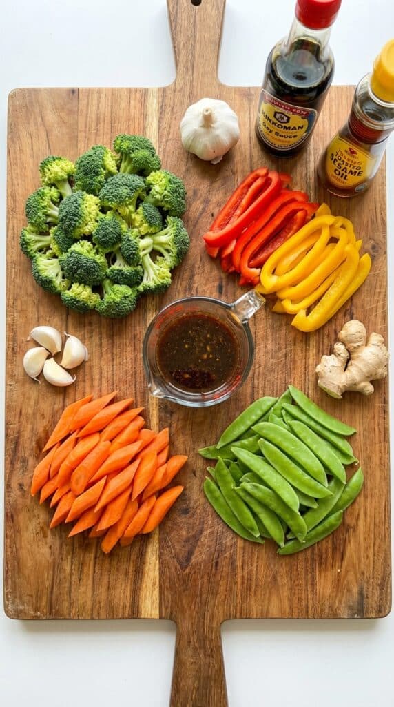 A flat lay showing neatly arranged piles of raw chopped vegetables, fresh ginger, garlic, and a bowl of stir-fry sauce on a wooden board.