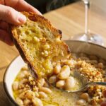 A close-up of crusty bread dipping into a bowl of garlic white beans with steam rising.