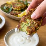 A close-up of a hand dipping a piece of zucchini fritter into a white sauce.