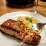 A close-up of a fork flaking a piece of cooked salmon, showing the moist pink interior and crispy skin, served with orzo.
