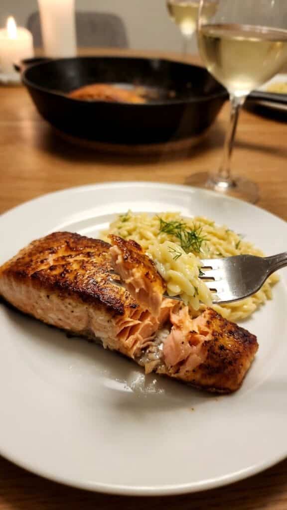 A close-up of a fork flaking a piece of cooked salmon, showing the moist pink interior and crispy skin, served with orzo.