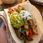 A close-up of a hand holding a loaded chicken fajita taco with guacamole and sour cream.