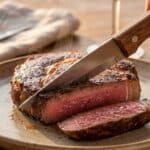 A close-up of a knife cutting into a ribeye steak revealing a juicy pink medium-rare interior.