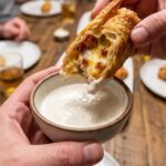 A close-up POV shot of a hand dipping a bitten jalapeño popper eggroll into ranch dressing.