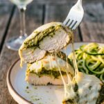 A close-up of a fork lifting a piece of pesto chicken with stringy melted cheese.
