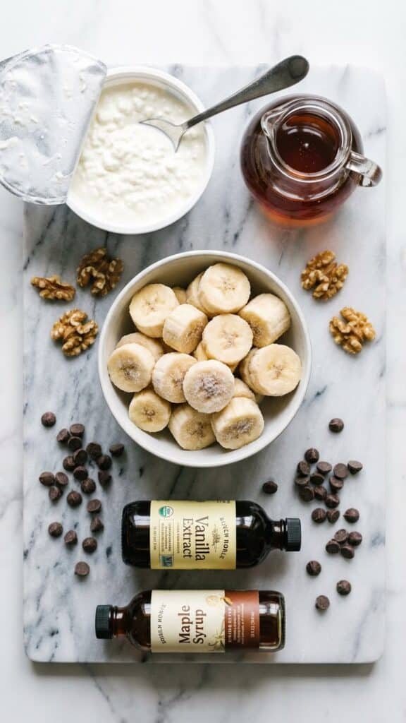 A flat lay showing frozen banana slices, a tub of cottage cheese, syrup, and toppings on a marble board.