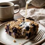 A close-up of milk being poured over a slice of baked blueberry oatmeal on a plate.