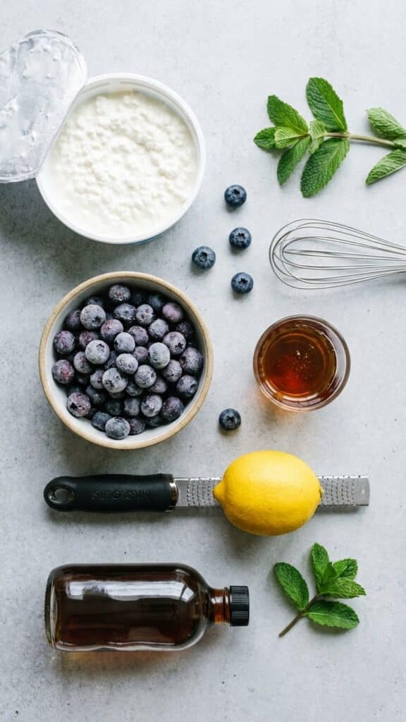 A flat lay showing cottage cheese, frozen blueberries, maple syrup, lemon, and vanilla.