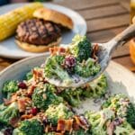 A close-up of a spoon lifting creamy broccoli salad with a burger plate in the background.