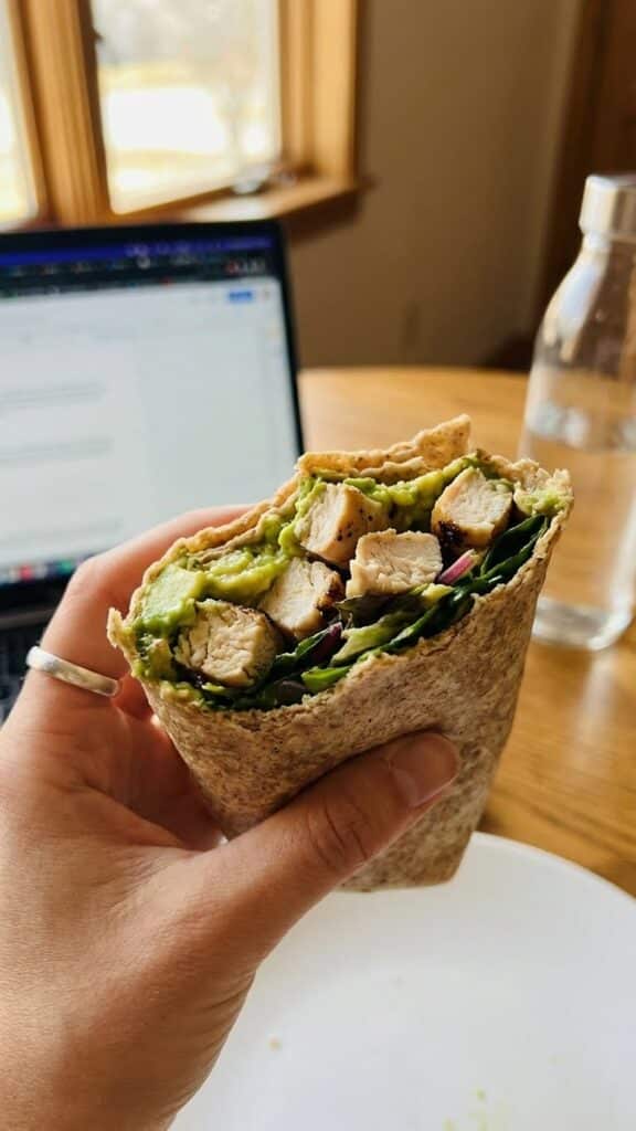 A close-up of a hand holding a chicken avocado wrap with a blurred laptop in the background.