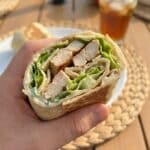 A close-up POV shot of a hand holding a fresh Chicken Caesar Wrap half, about to take a bite.