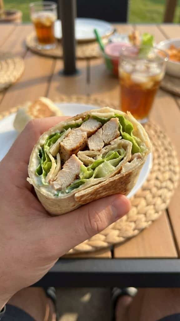 A close-up POV shot of a hand holding a fresh Chicken Caesar Wrap half, about to take a bite.