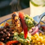 A close-up of a fork lifting a bite of chicken, peppers, and guacamole from a bowl.