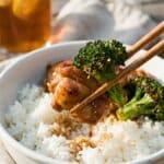A close-up of chopsticks lifting a piece of chicken and broccoli from a rice bowl.