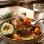 A close-up of a fork lifting a bite of chicken, sweet potato, and rice from a bowl with avocado.