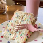 A close-up of a slice of cake showing rainbow sprinkles inside the white cake layers, with a fork taking a bite.
