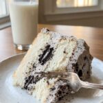 A close-up of a tall slice of Oreo cake on a plate with a fork taking a bite, and a glass of milk in the background.