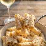 A close-up of a fork lifting rigatoni pasta coated in thick creamy sauce.