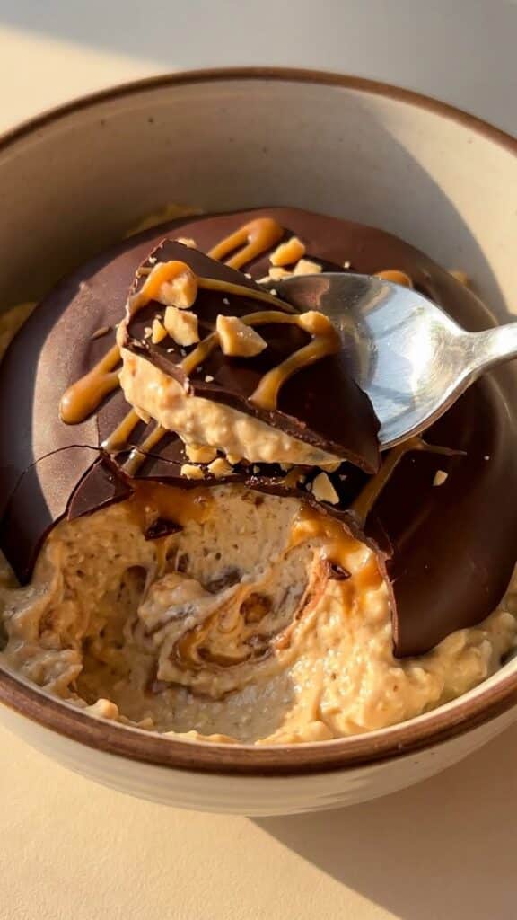 A close-up of a spoon cracking a hard chocolate shell to reveal smooth peanut butter filling.