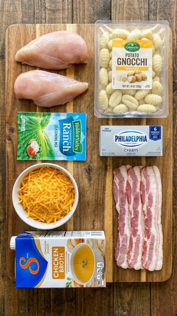 A flat lay showing raw chicken, gnocchi, ranch seasoning packet, cream cheese, cheddar, and bacon on a wooden board.