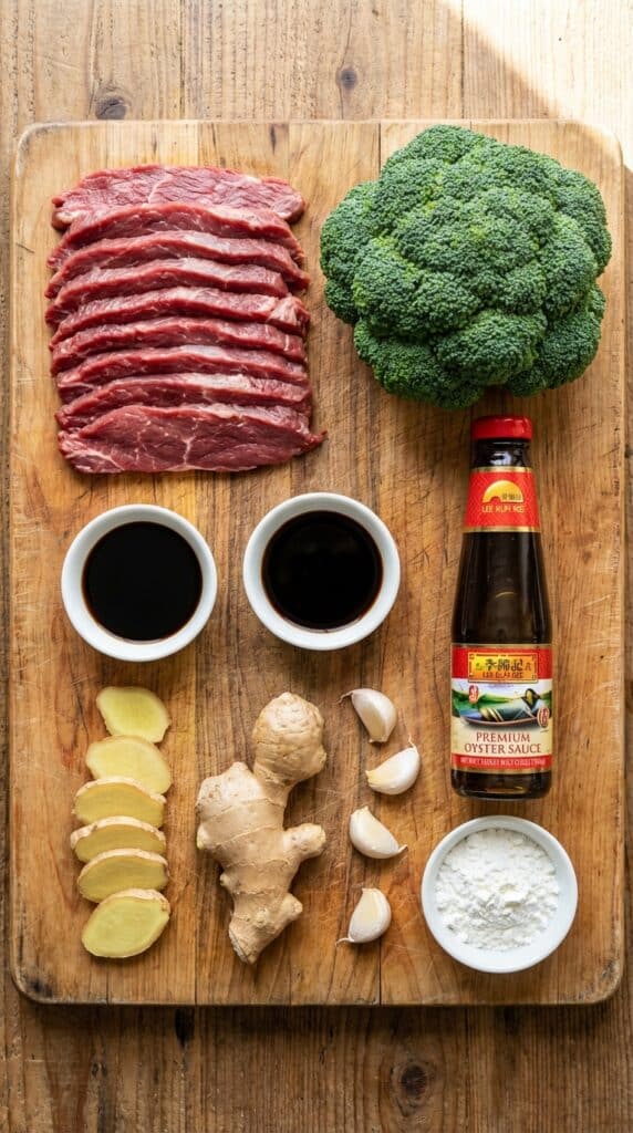 A flat lay showing sliced flank steak, broccoli florets, soy sauce, garlic, ginger, and cornstarch on a board.