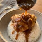 A close-up of a ladle pouring rich gravy over chicken and white rice in a bowl.