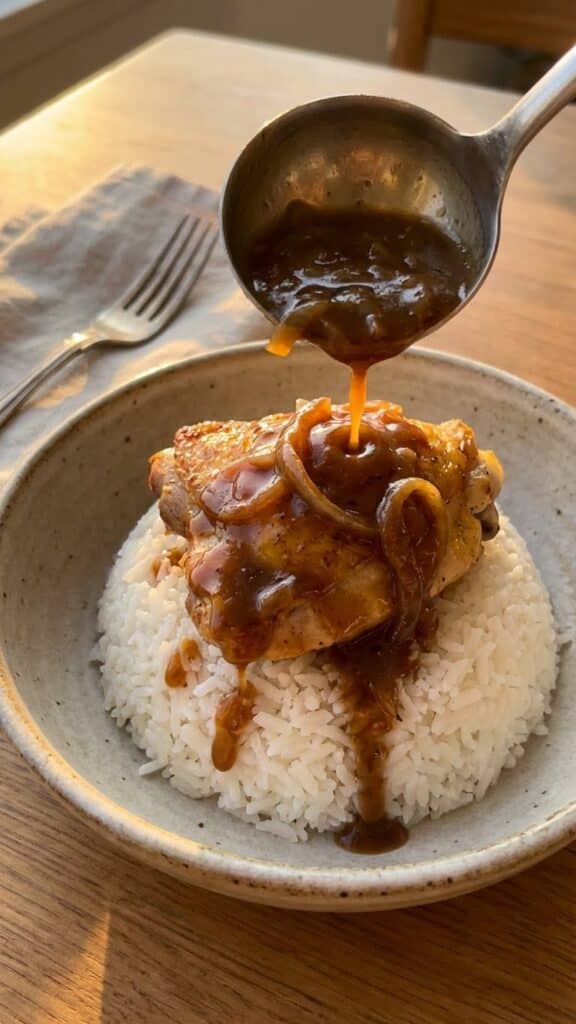 A close-up of a ladle pouring rich gravy over chicken and white rice in a bowl.