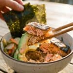 A close-up of chopsticks lifting a bite of salmon, rice, and avocado with spicy mayo.