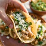 A close-up of a hand holding a street-style chicken taco topped with onion, cilantro, and cheese.