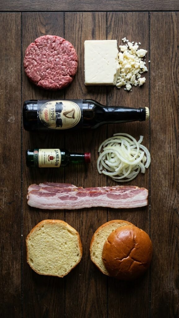 Overhead flat lay view of ingredients: raw beef, Irish cheddar cheese, dark stout beer, whiskey, onions, and bacon on a wooden table.