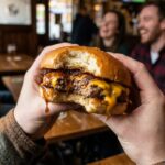 A close-up POV shot of hands holding a juicy Irish burger, with stout onions and cheese dripping out.
