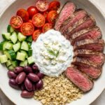 A top-down view of a healthy bowl with sliced steak, cucumbers, tomatoes, olives, feta, and tzatziki sauce.