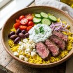 A close-up POV shot of a fork lifting a mixed bite of steak, vegetables, feta, and tzatziki from a bowl.