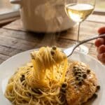 A close-up of a fork lifting a piece of lemon chicken and angel hair pasta from a plate.