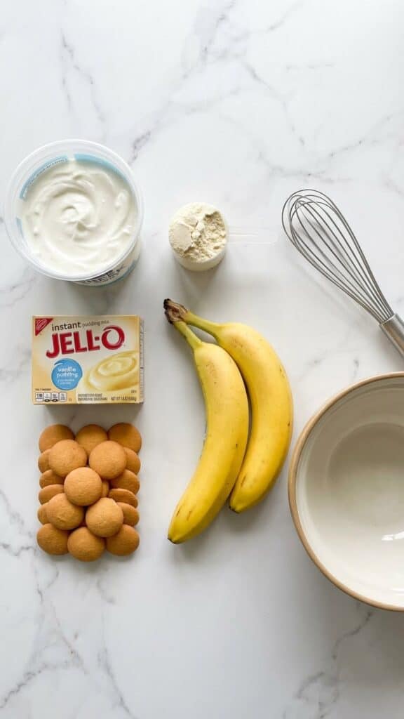 A flat lay showing Greek yogurt, protein powder, bananas, pudding mix, and Nilla wafers on a marble table.
