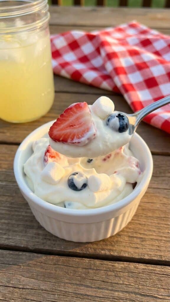 An overhead flat lay of ingredients including cream cheese, cool whip, strawberries, blueberries, and marshmallows on a marble surface.